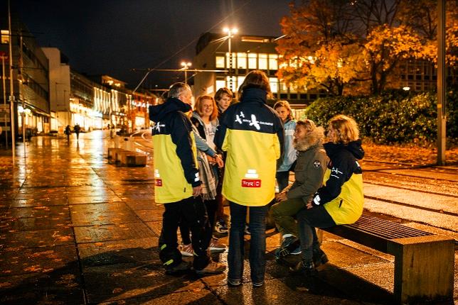 En gruppe mennesker med Ntteravn-jakker som ler og snakker sammen  ved en benk på kvelden