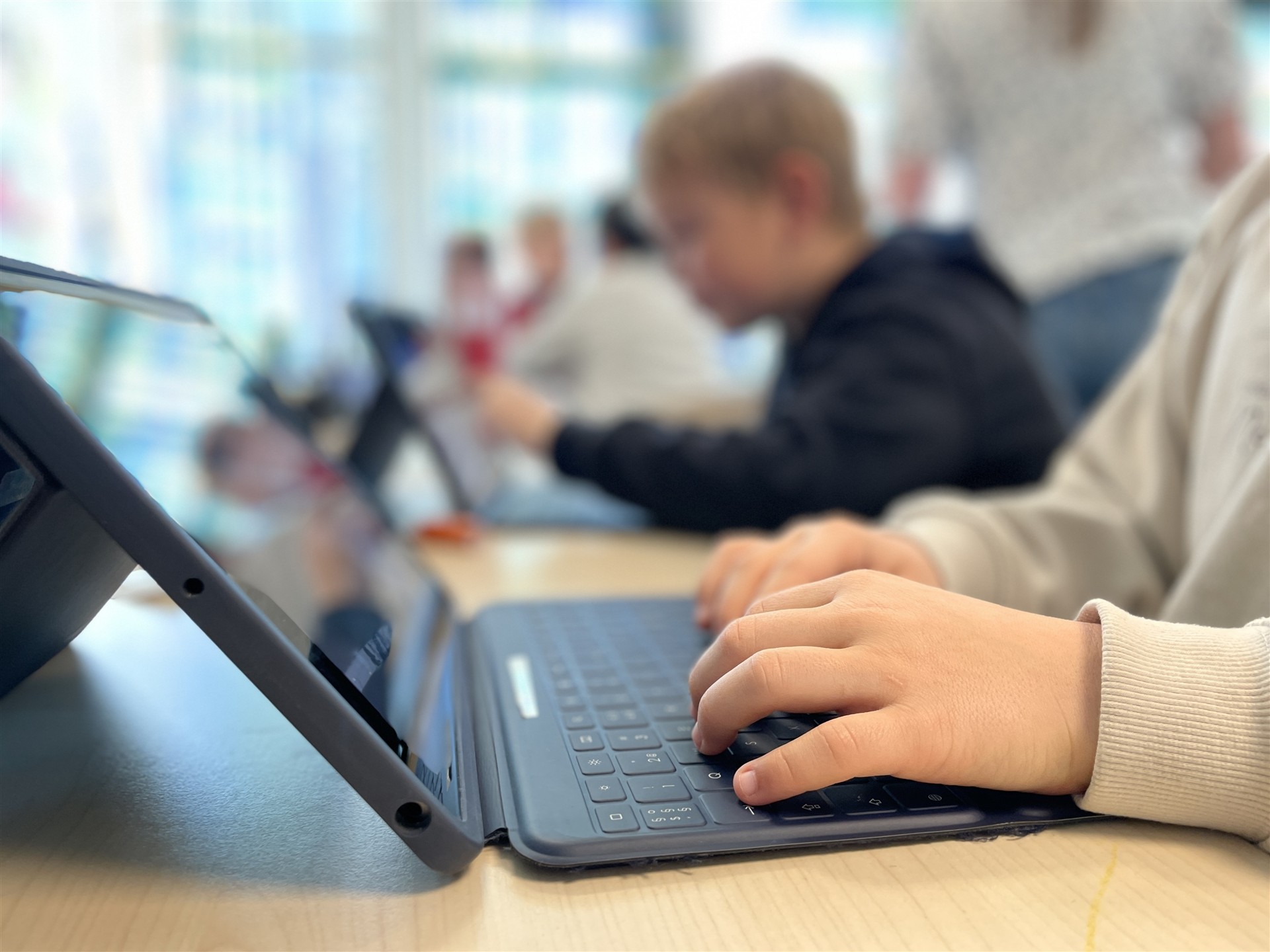 Barnehender på en laptop - og elever i bakgrunnen som er i defokus